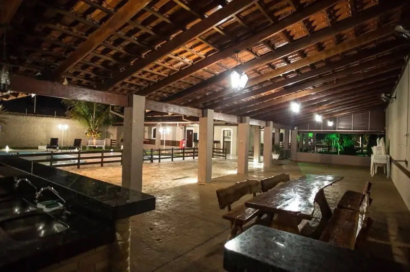 Área gourmet à noite, com bancada de granito, mesas de madeira e a piscina iluminada ao fundo
