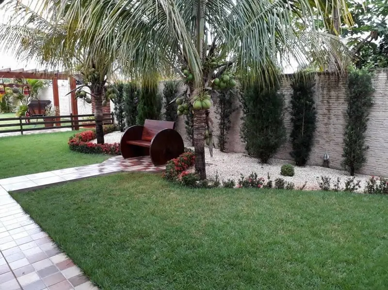 Jardim com gramado, coqueiros, canteiro de flores e banco decorativo vermelho