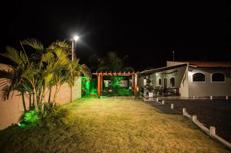 Jardim iluminado à noite, com palmeiras, pérgola e a casa ao fundo