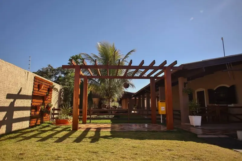 Pérgola de madeira sobre o gramado, com a varanda da casa ao fundo
