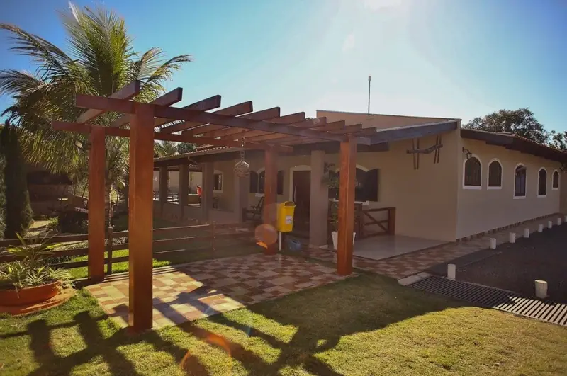 Pérgola de madeira no jardim, com gramado, coqueiro e a casa ao fundo