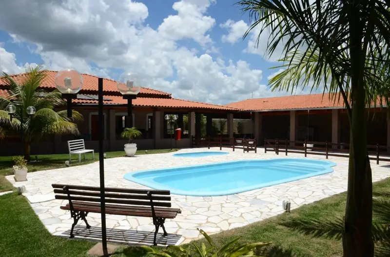 Piscina da Chácara Império com banco de madeira, palmeira e a casa ao fundo, em dia de céu azul