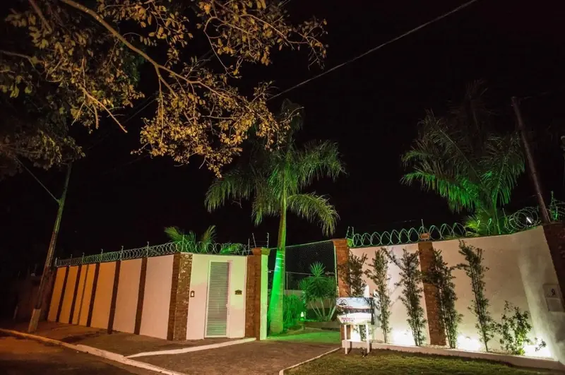 Portão de entrada da Chácara Império iluminado à noite, com palmeiras e muro com trepadeiras